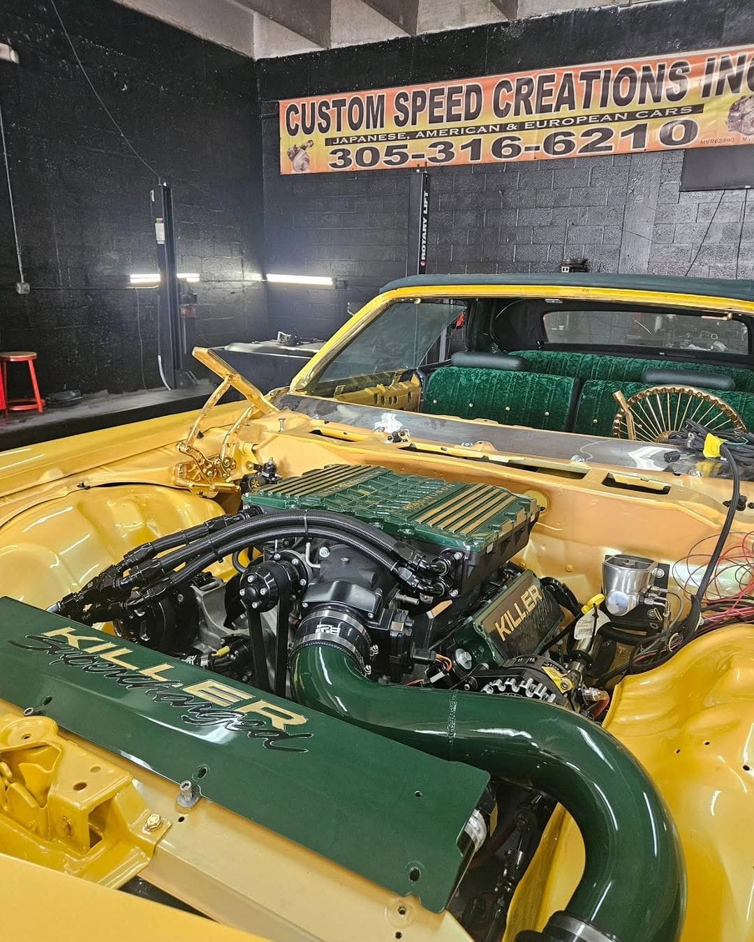 The engine bay of a yellow classic convertible with a green KILLER intake, in the Custom Speed Creations shop