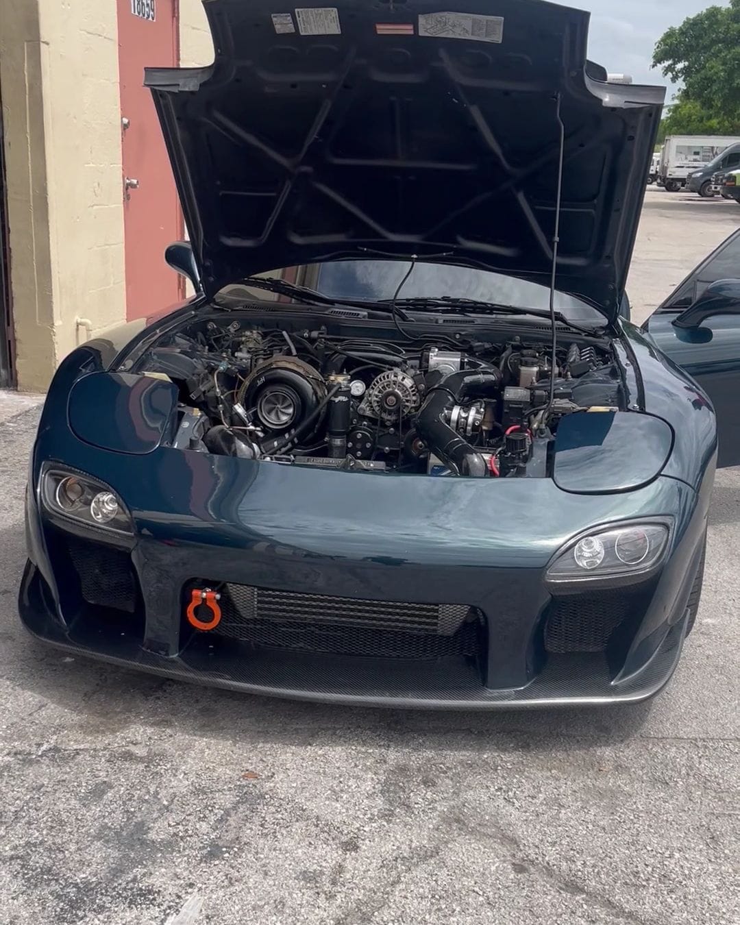 A dark green Mazda RX-7 with the hood up, showing its turbocharged engine, outside the shop